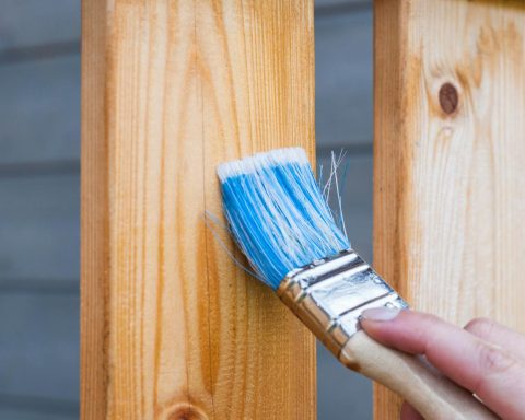 A hand painting a wooden fence plank with a blue paintbrush outdoors. Perfect for DIY and renovation themes.